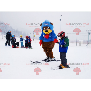 Brązowy i żółty niedźwiedź maskotka w stroju narciarskim.